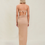 A woman with short blonde hair stands facing away, wearing a tan strapless top tied at the back and the Gwen Skirt, which features a high slit. The background is plain and light-colored.