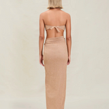 A woman with short blonde hair stands facing away, wearing a tan strapless top tied at the back and the Gwen Skirt, which features a high slit. The background is plain and light-colored.