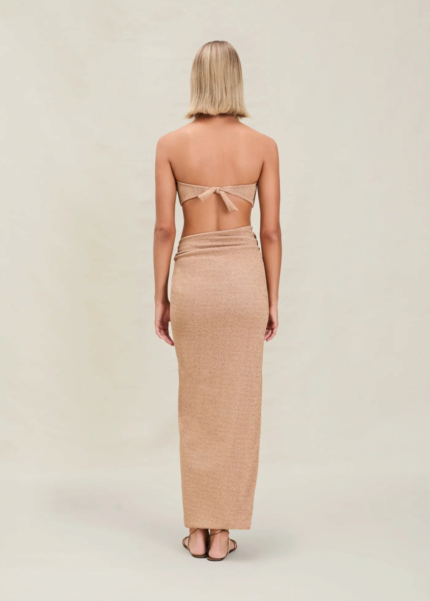 A woman with short blonde hair stands facing away, wearing a tan strapless top tied at the back and the Gwen Skirt, which features a high slit. The background is plain and light-colored.