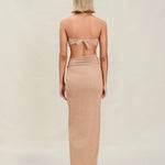 A woman with short blonde hair stands facing away, wearing a tan strapless top tied at the back and the Gwen Skirt, which features a high slit. The background is plain and light-colored.