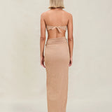 A woman with short blonde hair stands facing away, wearing a tan strapless top tied at the back and the Gwen Skirt, which features a high slit. The background is plain and light-colored.