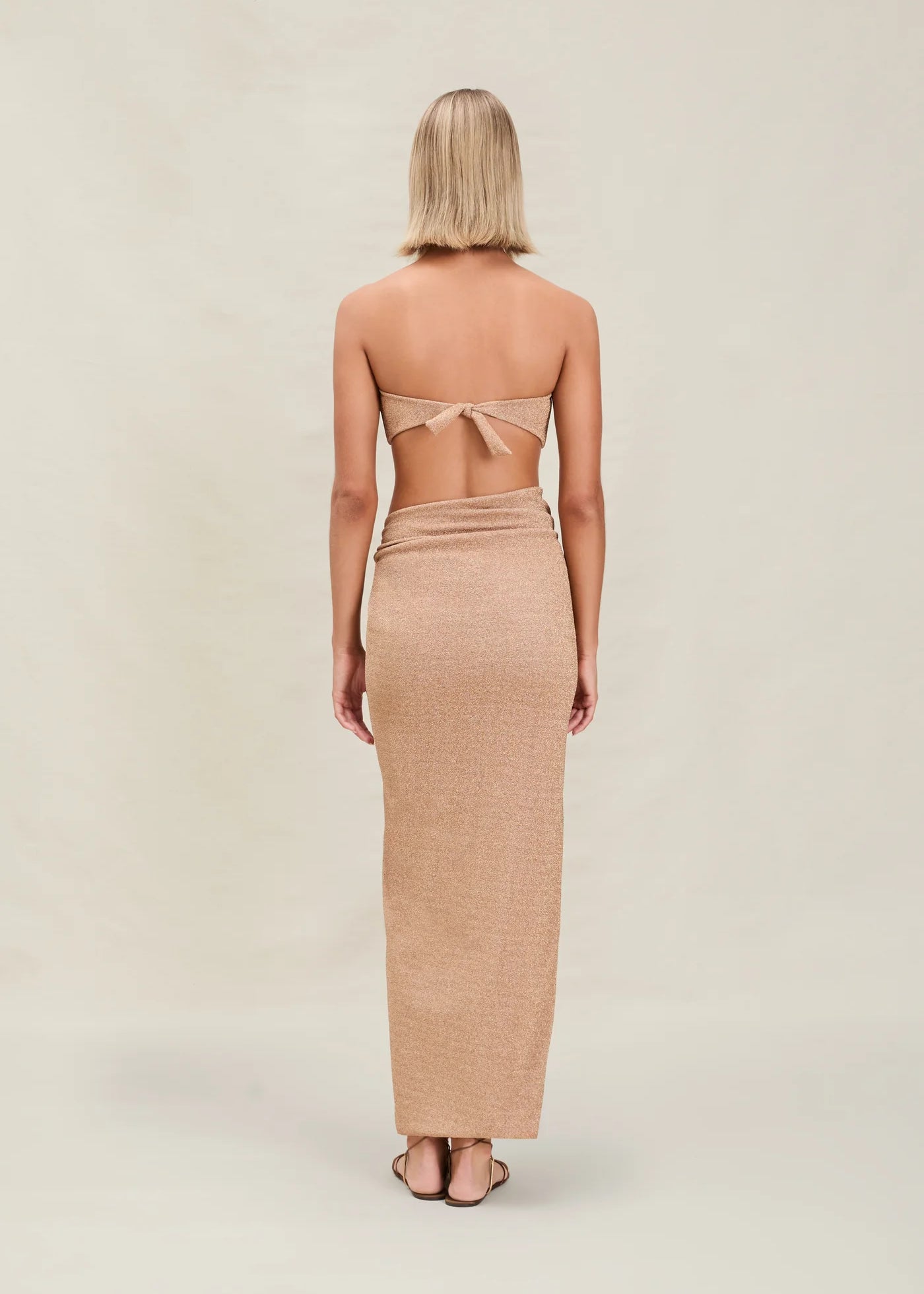 A woman with short blonde hair stands facing away, wearing a tan strapless top tied at the back and the Gwen Skirt, which features a high slit. The background is plain and light-colored.