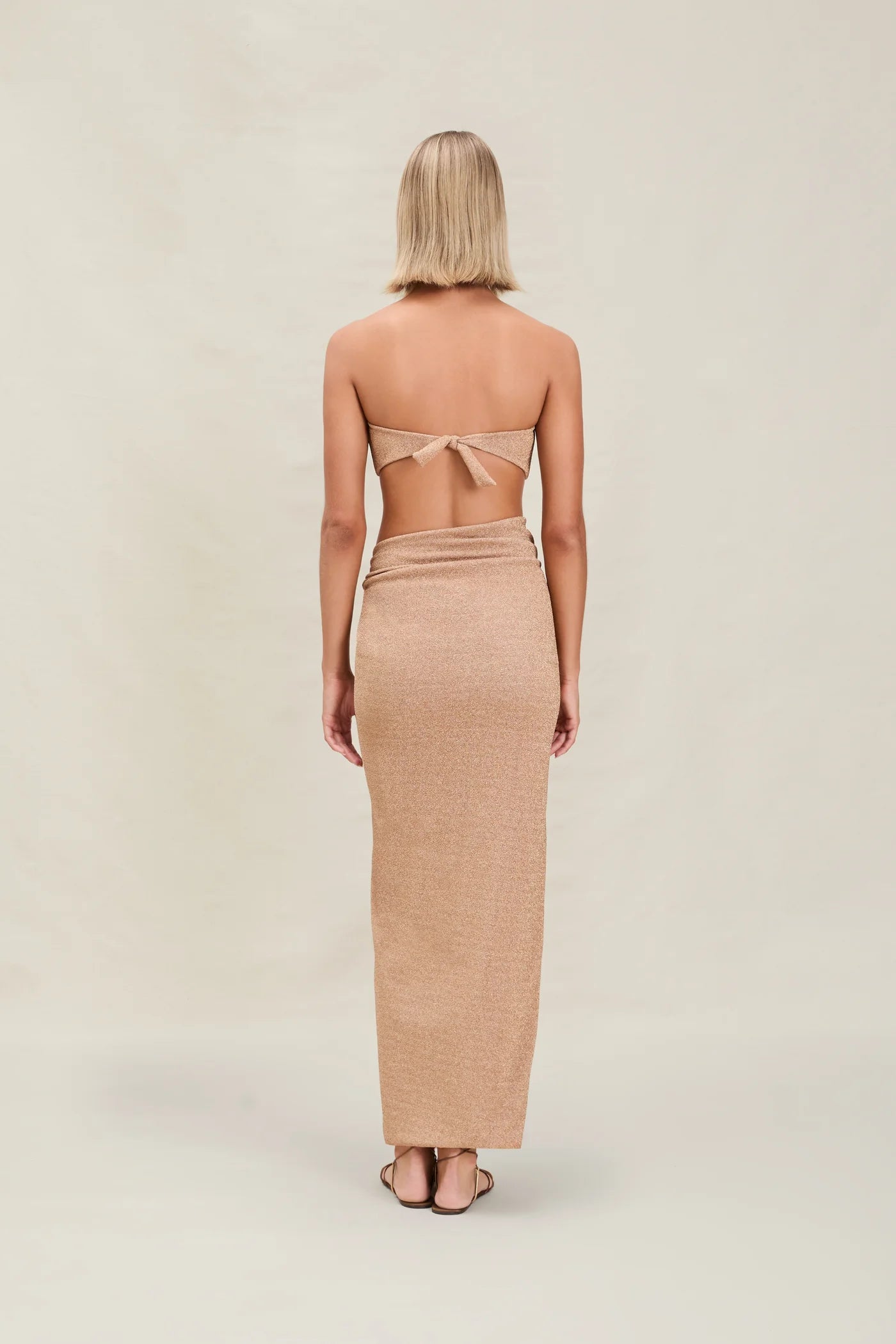 A woman with short blonde hair stands facing away, wearing a tan strapless top tied at the back and the Gwen Skirt, which features a high slit. The background is plain and light-colored.