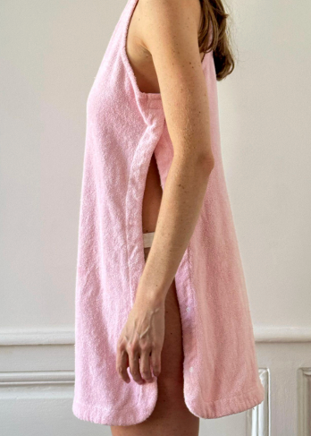 A woman stands sideways wearing the Cruise Dress, a sleeveless, light pink towel-style garment with open sides that reveal part of her underwear. The background is a plain, light-colored wall with white trim.