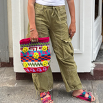 A person wearing olive cargo pants, a white top, and colorful sandals stands on a sidewalk, holding the Smileys Bag—a handmade crocheted tote adorned with smiley faces and bright geometric patterns.