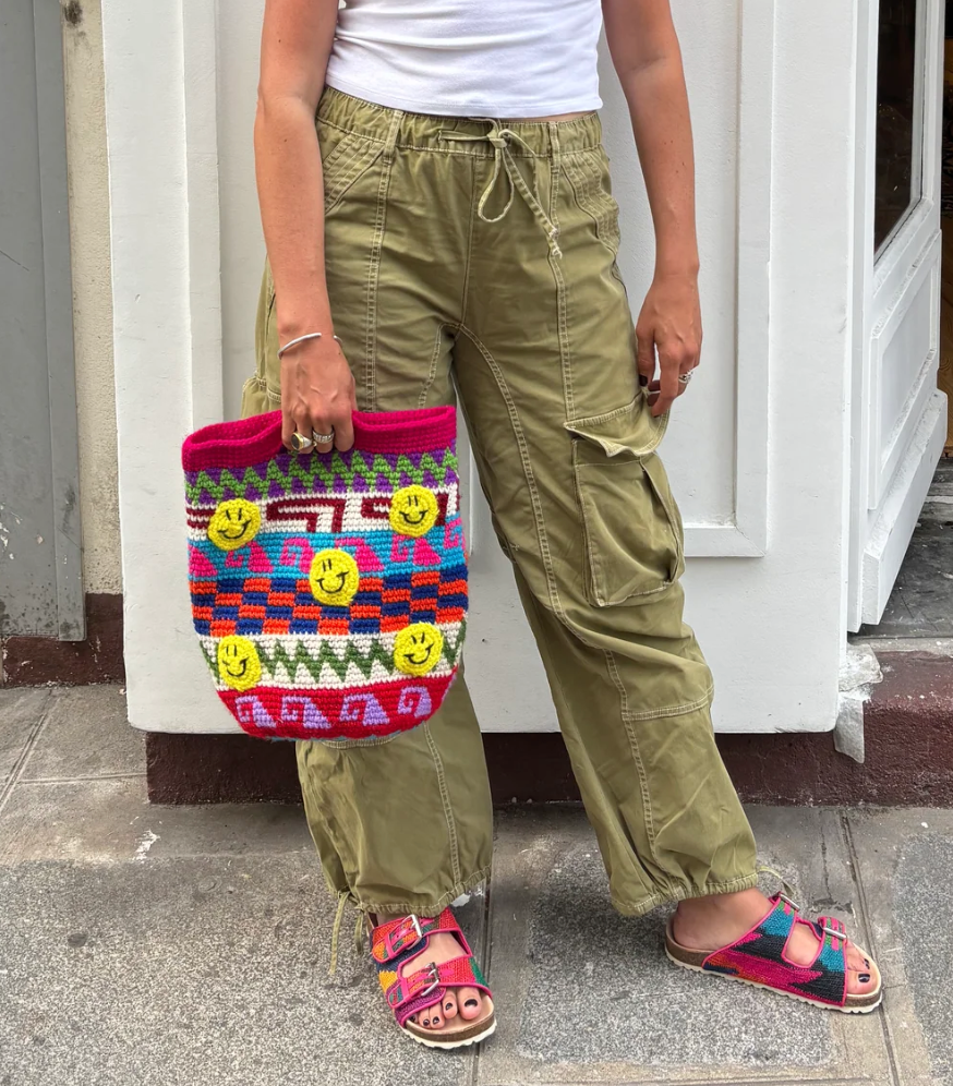A person wearing olive cargo pants, a white top, and colorful sandals stands on a sidewalk, holding the Smileys Bag—a handmade crocheted tote adorned with smiley faces and bright geometric patterns.