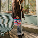 A person in a brown jacket and jeans stands by a bench, holding the Champinones Bag—a colorful zigzag-patterned handwoven accessory featuring a mushroom design. Green plants are visible through the glass wall behind them.