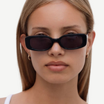 A woman with straight, light brown hair wears the Solene slim rectangle sunglasses in black, which provide 100% UV protection, paired with gold hoop earrings. She is looking straight ahead with a neutral expression and dressed in a white top against a plain background.