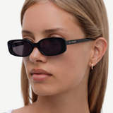 A woman with straight, light brown hair wears the Solene black slim rectangle sunglasses, which offer 100% UV protection, along with a small gold hoop earring. She is looking to the side with a neutral expression, dressed in a white top against a plain background.