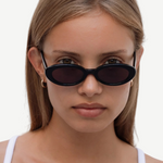 A young woman with straight light brown hair wears the Sylvie sunglasses in classic black and a white tank top, looking directly at the camera against a plain white background.