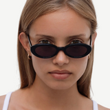 A young woman with straight light brown hair wears the Sylvie sunglasses in classic black and a white tank top, looking directly at the camera against a plain white background.