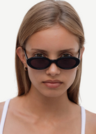 A young woman with straight light brown hair wears the Sylvie sunglasses in classic black and a white tank top, looking directly at the camera against a plain white background.