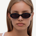 A person with straight, light brown hair wears the Sylvie sunglasses in black, paired with small hoop earrings and a white top, looking directly at the camera against a plain light background.