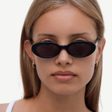 A person with straight, light brown hair wears the Sylvie sunglasses in black, paired with small hoop earrings and a white top, looking directly at the camera against a plain light background.