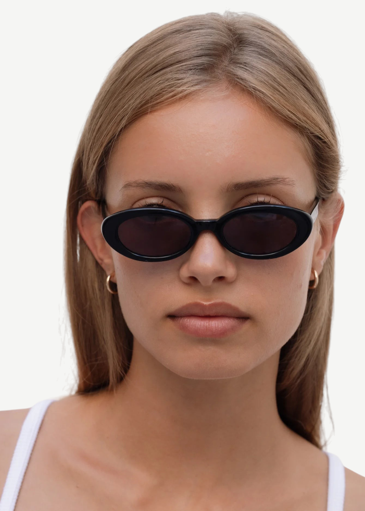 A person with straight, light brown hair wears the Sylvie sunglasses in black, paired with small hoop earrings and a white top, looking directly at the camera against a plain light background.