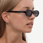 A woman with straight, light brown hair wears the Sylvie sunglasses in black, paired with a gold hoop earring and a white tank top—her effortlessly chic look enhanced as she glances to the side against a plain, light background.