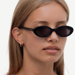 A person with straight, light brown hair is wearing the Sylvie black oval sunglasses with “UN-DONE” text on the arm and a white top, standing against a plain white background.