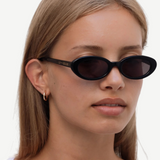 A person with straight, light brown hair is wearing the Sylvie black oval sunglasses with “UN-DONE” text on the arm and a white top, standing against a plain white background.