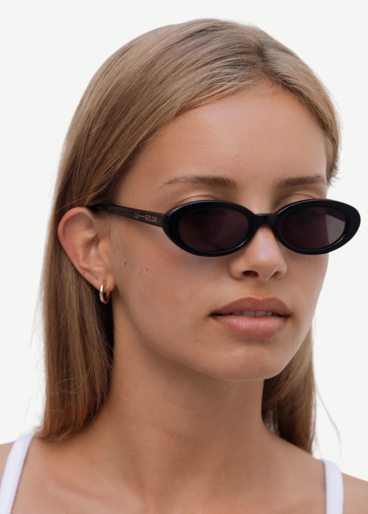 A person with straight, light brown hair is wearing the Sylvie black oval sunglasses with “UN-DONE” text on the arm and a white top, standing against a plain white background.