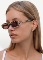 A woman with straight, light brown hair wears Sylvie oval tortoiseshell sunglasses with UV400 protection and a small gold hoop earring. She has a neutral expression and is dressed in a white top against a plain white background.