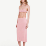 A woman stands against a plain light background, wearing a light pink bralette and the Tube Skirt crafted from soft bamboo fabric, paired with black slip-on shoes. Her hair is loose, and she has a neutral expression, highlighting the minimalist design of her outfit.