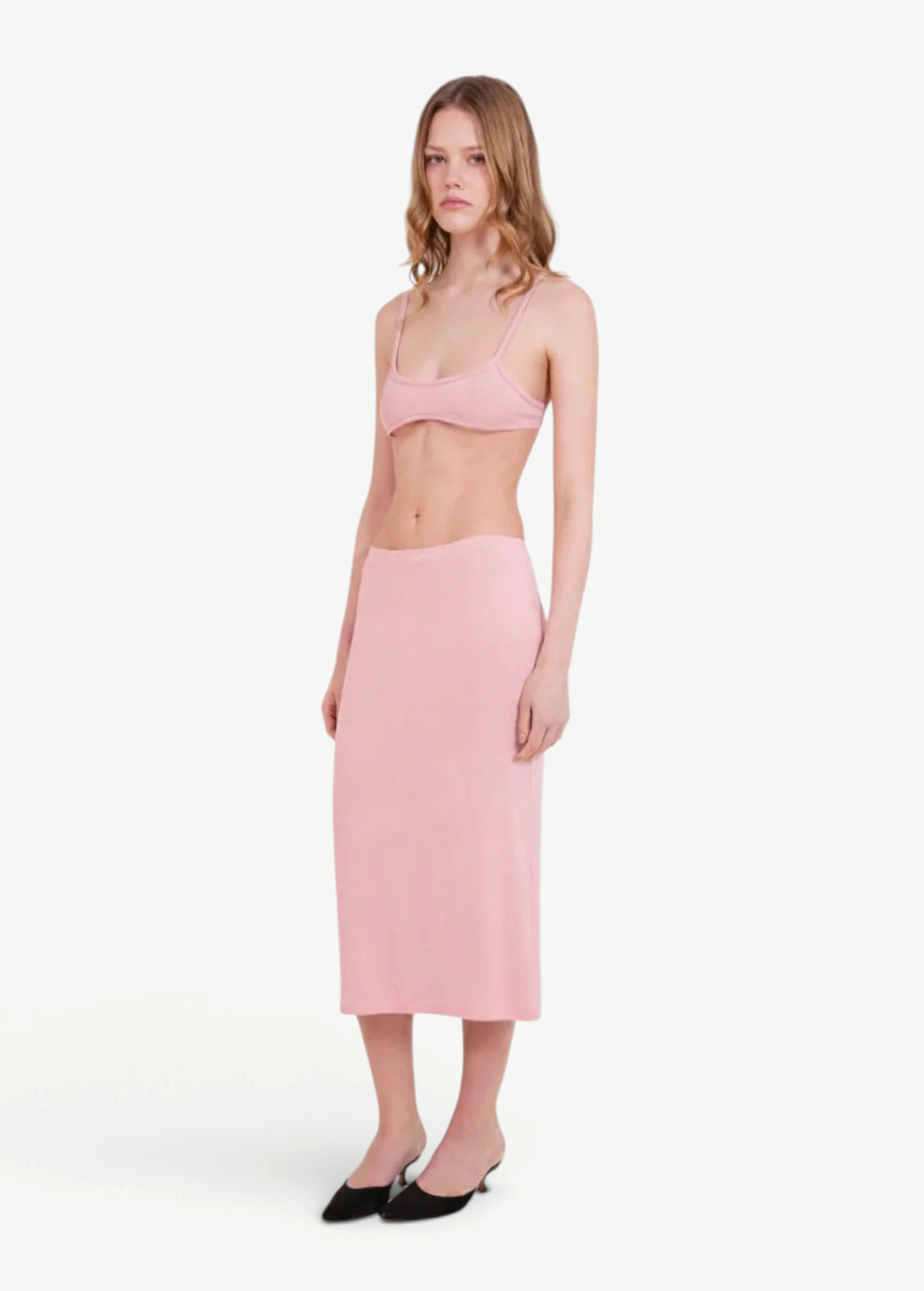 A woman stands against a plain light background, wearing a light pink bralette and the Tube Skirt crafted from soft bamboo fabric, paired with black slip-on shoes. Her hair is loose, and she has a neutral expression, highlighting the minimalist design of her outfit.