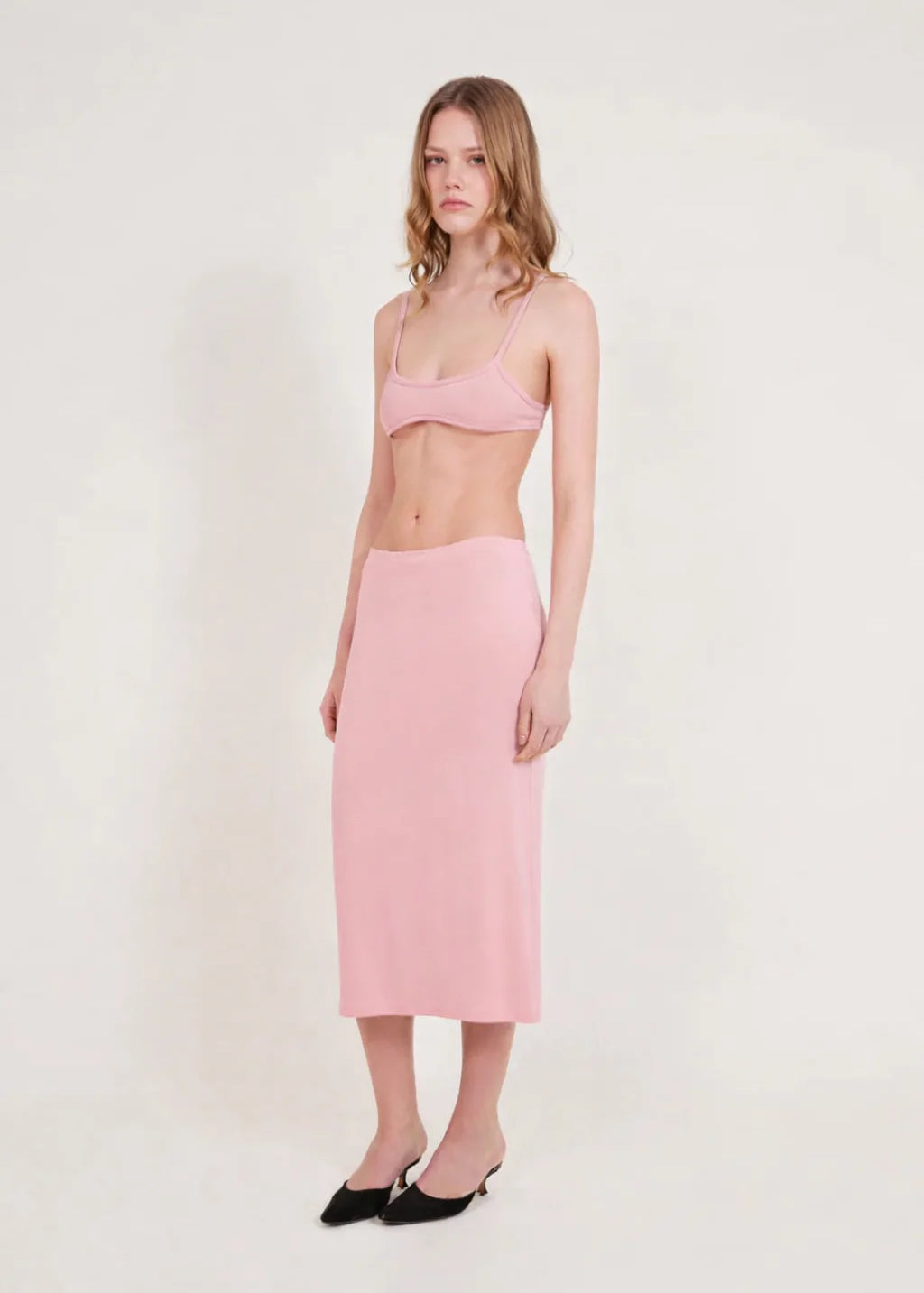 A woman stands against a plain light background, wearing a light pink bralette and the Tube Skirt crafted from soft bamboo fabric, paired with black slip-on shoes. Her hair is loose, and she has a neutral expression, highlighting the minimalist design of her outfit.