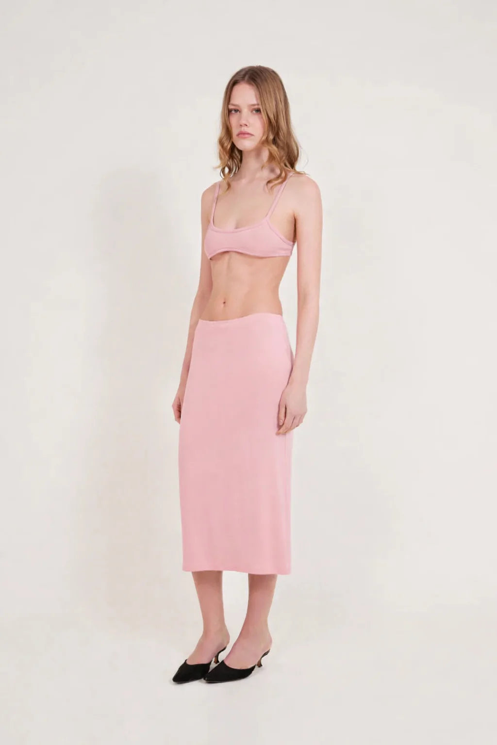 A woman stands against a plain light background, wearing a light pink bralette and the Tube Skirt crafted from soft bamboo fabric, paired with black slip-on shoes. Her hair is loose, and she has a neutral expression, highlighting the minimalist design of her outfit.