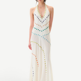 A person stands against a plain white background wearing the Vicka Dress—a sleeveless, floor-length white backless dress embellished with colorful circular beads arranged in curved lines, featuring intricate bead embroidery and a subtly flared hemline.