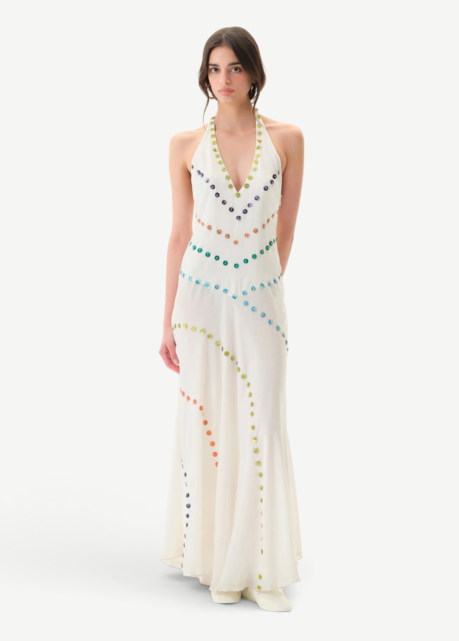 A person stands against a plain white background wearing the Vicka Dress—a sleeveless, floor-length white backless dress embellished with colorful circular beads arranged in curved lines, featuring intricate bead embroidery and a subtly flared hemline.