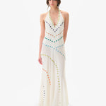 A person stands against a plain white background wearing the Vicka Dress—a sleeveless, floor-length white backless dress embellished with colorful circular beads arranged in curved lines, featuring intricate bead embroidery and a subtly flared hemline.