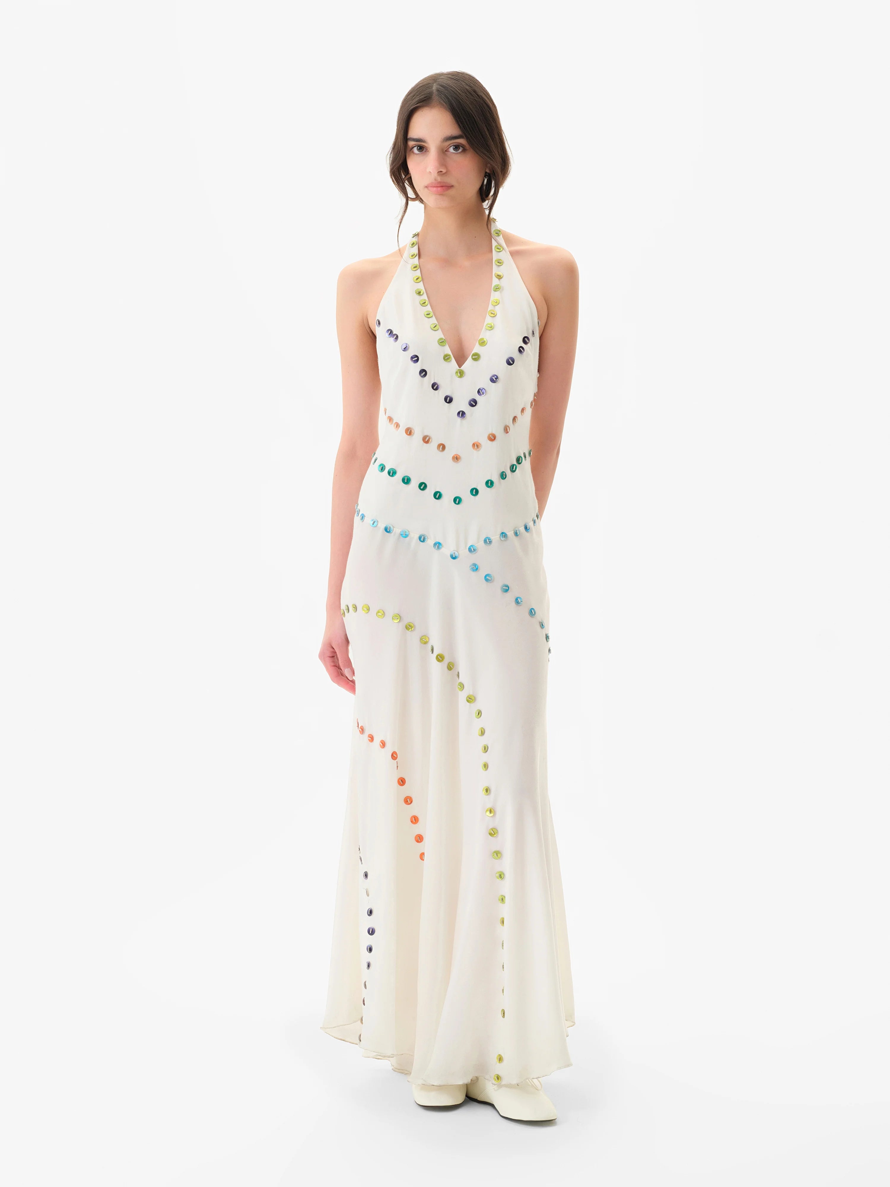 A person stands against a plain white background wearing the Vicka Dress—a sleeveless, floor-length white backless dress embellished with colorful circular beads arranged in curved lines, featuring intricate bead embroidery and a subtly flared hemline.