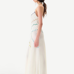 Woman wearing the Vicka Dress—a long, sleeveless white backless dress featuring colorful bead embroidery—standing sideways against a plain white background. She has long brown hair styled in a low ponytail and is paired with white shoes.