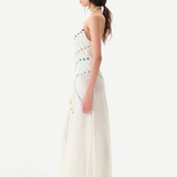 Woman wearing the Vicka Dress—a long, sleeveless white backless dress featuring colorful bead embroidery—standing sideways against a plain white background. She has long brown hair styled in a low ponytail and is paired with white shoes.