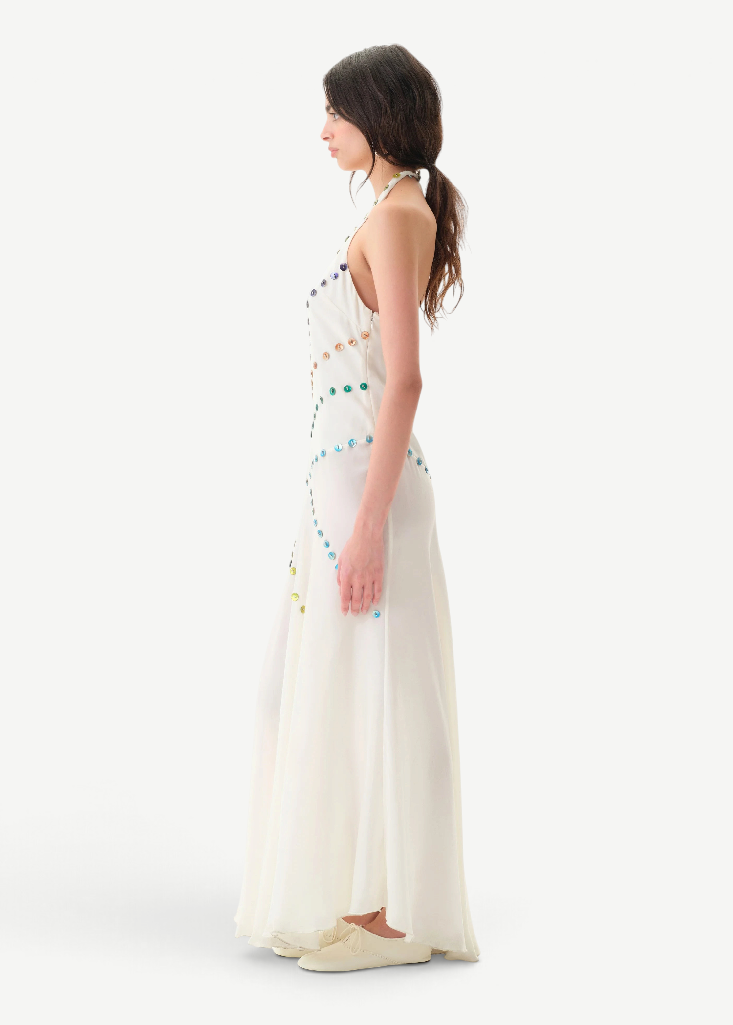 Woman wearing the Vicka Dress—a long, sleeveless white backless dress featuring colorful bead embroidery—standing sideways against a plain white background. She has long brown hair styled in a low ponytail and is paired with white shoes.
