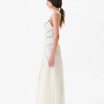 Woman wearing the Vicka Dress—a long, sleeveless white backless dress featuring colorful bead embroidery—standing sideways against a plain white background. She has long brown hair styled in a low ponytail and is paired with white shoes.