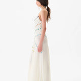 Woman wearing the Vicka Dress—a long, sleeveless white backless dress featuring colorful bead embroidery—standing sideways against a plain white background. She has long brown hair styled in a low ponytail and is paired with white shoes.