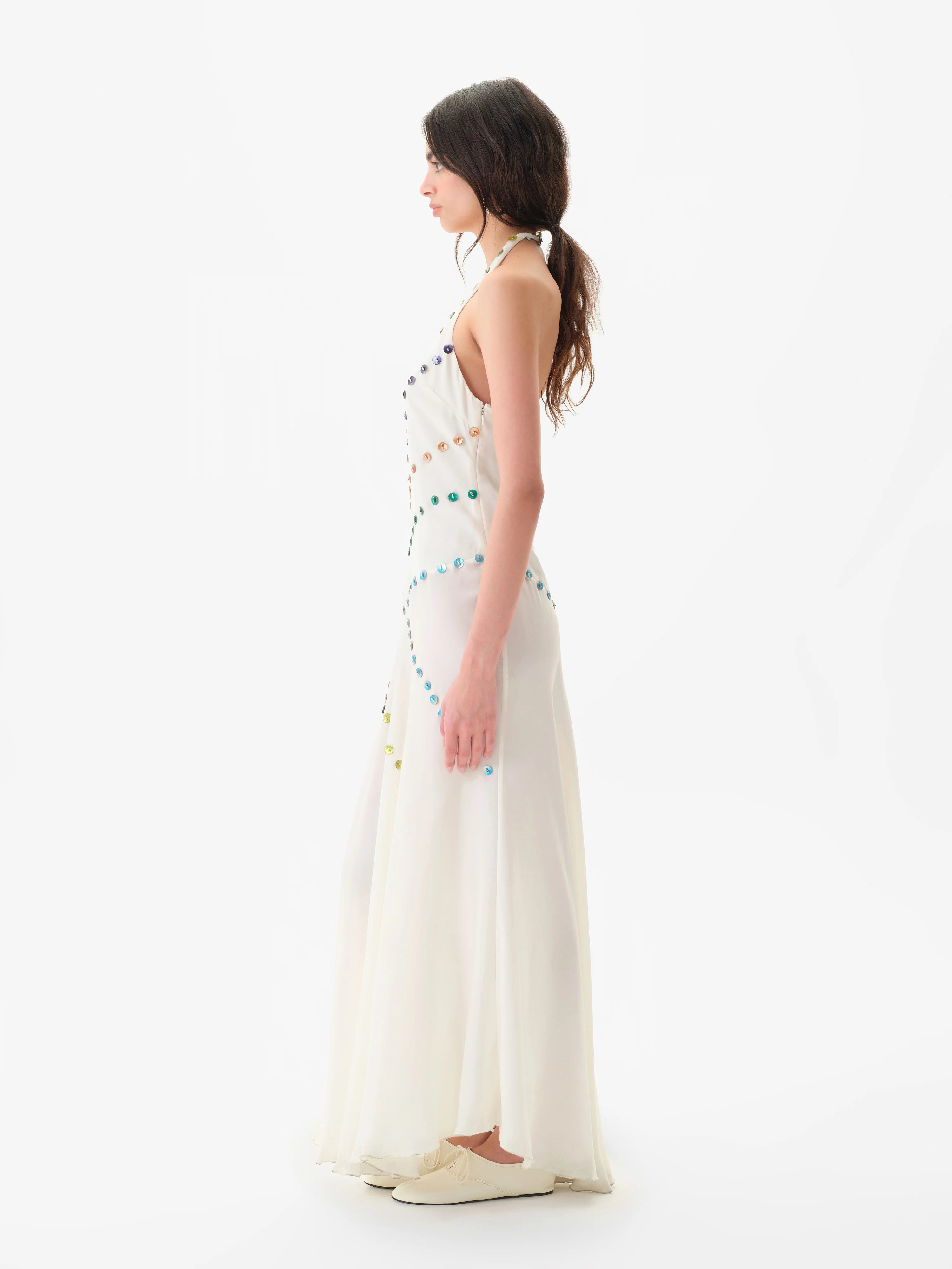 Woman wearing the Vicka Dress—a long, sleeveless white backless dress featuring colorful bead embroidery—standing sideways against a plain white background. She has long brown hair styled in a low ponytail and is paired with white shoes.