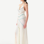 A woman with dark hair in a ponytail, wearing white shoes, stands sideways against a plain white background in the Vicka Dress—a long, white halter backless style featuring diagonal rows of colorful buttons and a gracefully flared hemline.