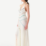 A woman with dark hair in a ponytail, wearing white shoes, stands sideways against a plain white background in the Vicka Dress—a long, white halter backless style featuring diagonal rows of colorful buttons and a gracefully flared hemline.