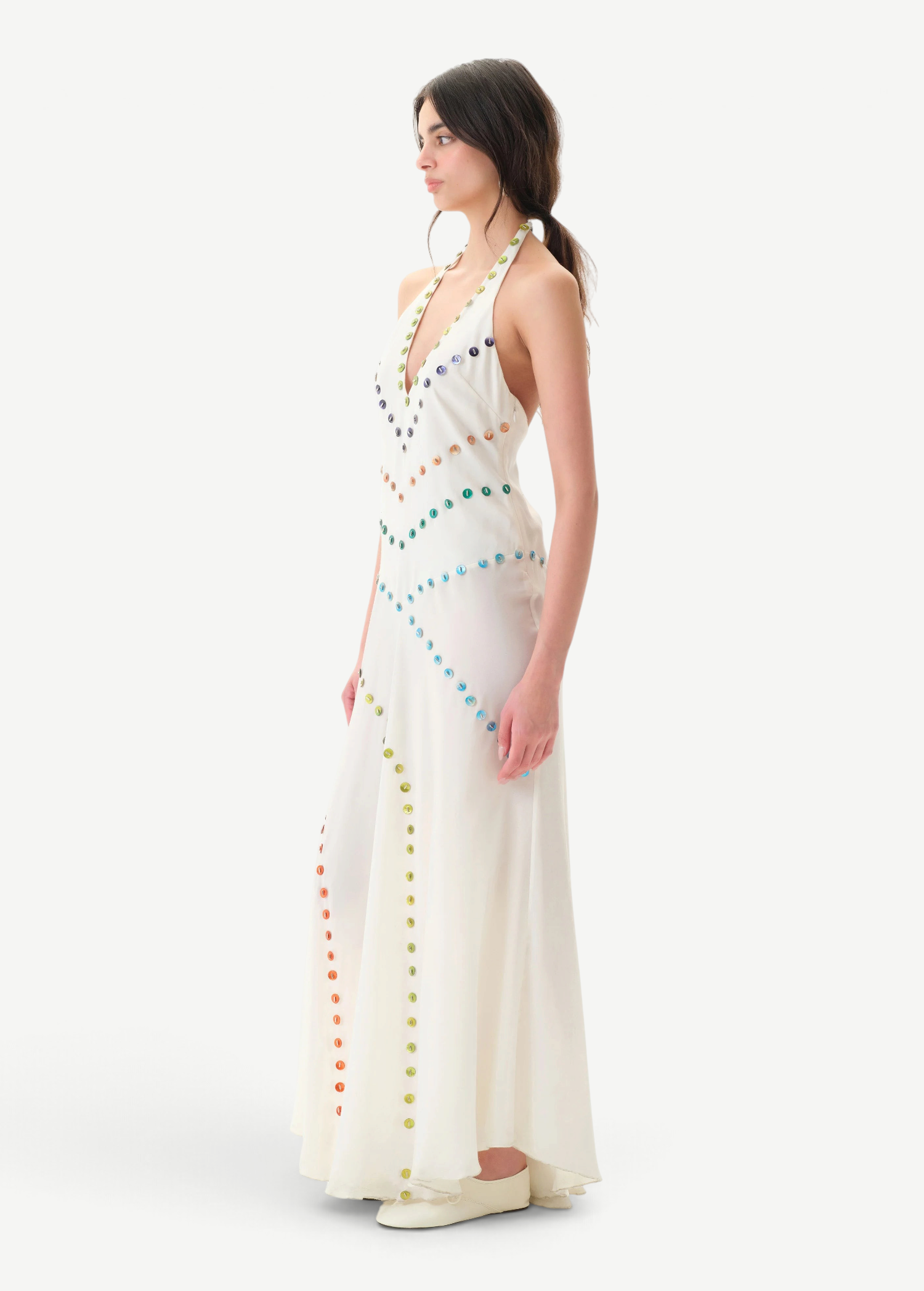 A woman with dark hair in a ponytail, wearing white shoes, stands sideways against a plain white background in the Vicka Dress—a long, white halter backless style featuring diagonal rows of colorful buttons and a gracefully flared hemline.