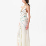A woman with dark hair in a ponytail, wearing white shoes, stands sideways against a plain white background in the Vicka Dress—a long, white halter backless style featuring diagonal rows of colorful buttons and a gracefully flared hemline.
