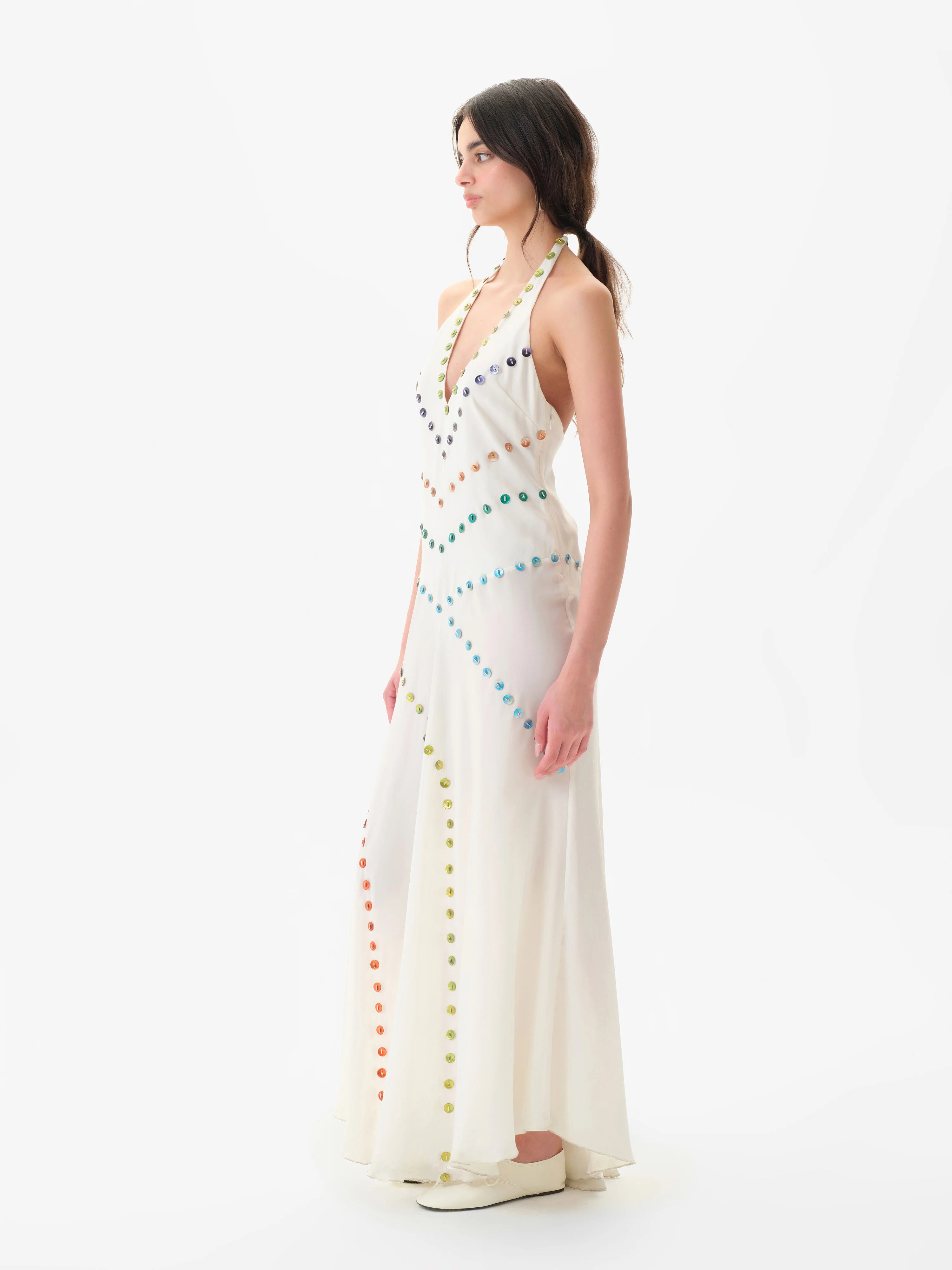 A woman with dark hair in a ponytail, wearing white shoes, stands sideways against a plain white background in the Vicka Dress—a long, white halter backless style featuring diagonal rows of colorful buttons and a gracefully flared hemline.