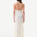 A woman with long brown hair stands facing away, wearing the Vicka Dress—a backless, floor-length white gown featuring a flared hemline and a beaded blue accent along the lower back. Elegant bead embroidery elevates the minimalist design. The background is plain white.