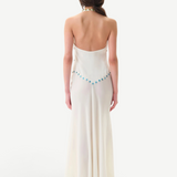 A woman with long brown hair stands facing away, wearing the Vicka Dress—a backless, floor-length white gown featuring a flared hemline and a beaded blue accent along the lower back. Elegant bead embroidery elevates the minimalist design. The background is plain white.
