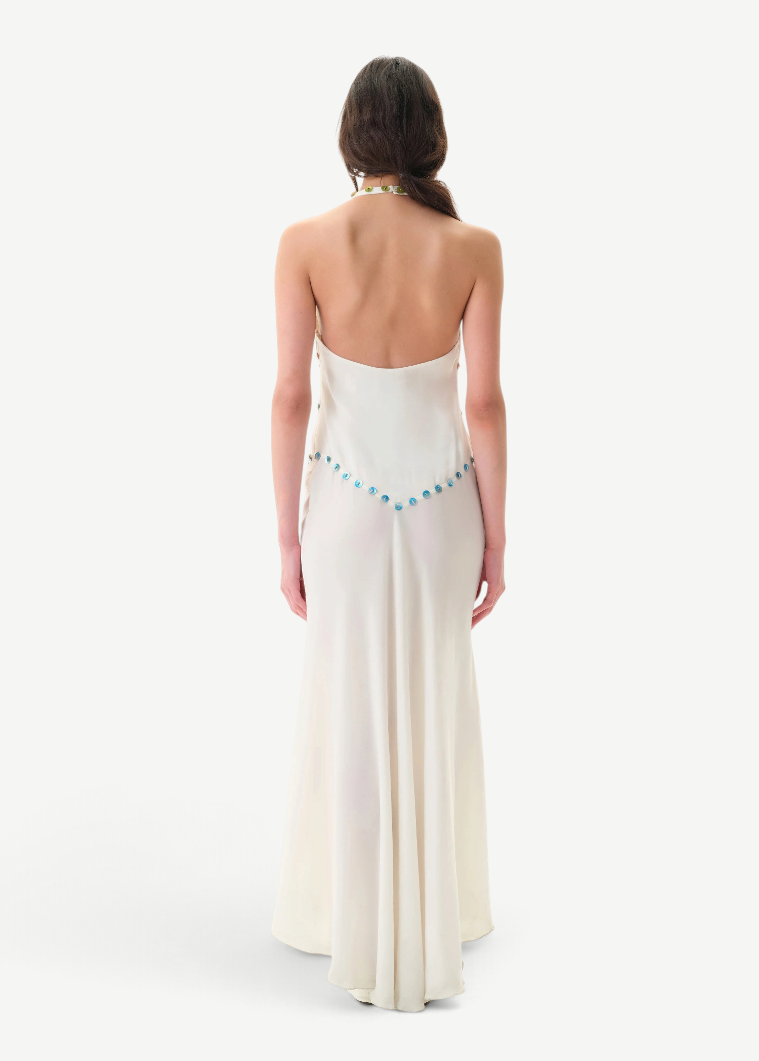A woman with long brown hair stands facing away, wearing the Vicka Dress—a backless, floor-length white gown featuring a flared hemline and a beaded blue accent along the lower back. Elegant bead embroidery elevates the minimalist design. The background is plain white.
