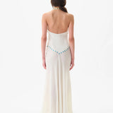 A woman with long brown hair stands facing away, wearing the Vicka Dress—a backless, floor-length white gown featuring a flared hemline and a beaded blue accent along the lower back. Elegant bead embroidery elevates the minimalist design. The background is plain white.