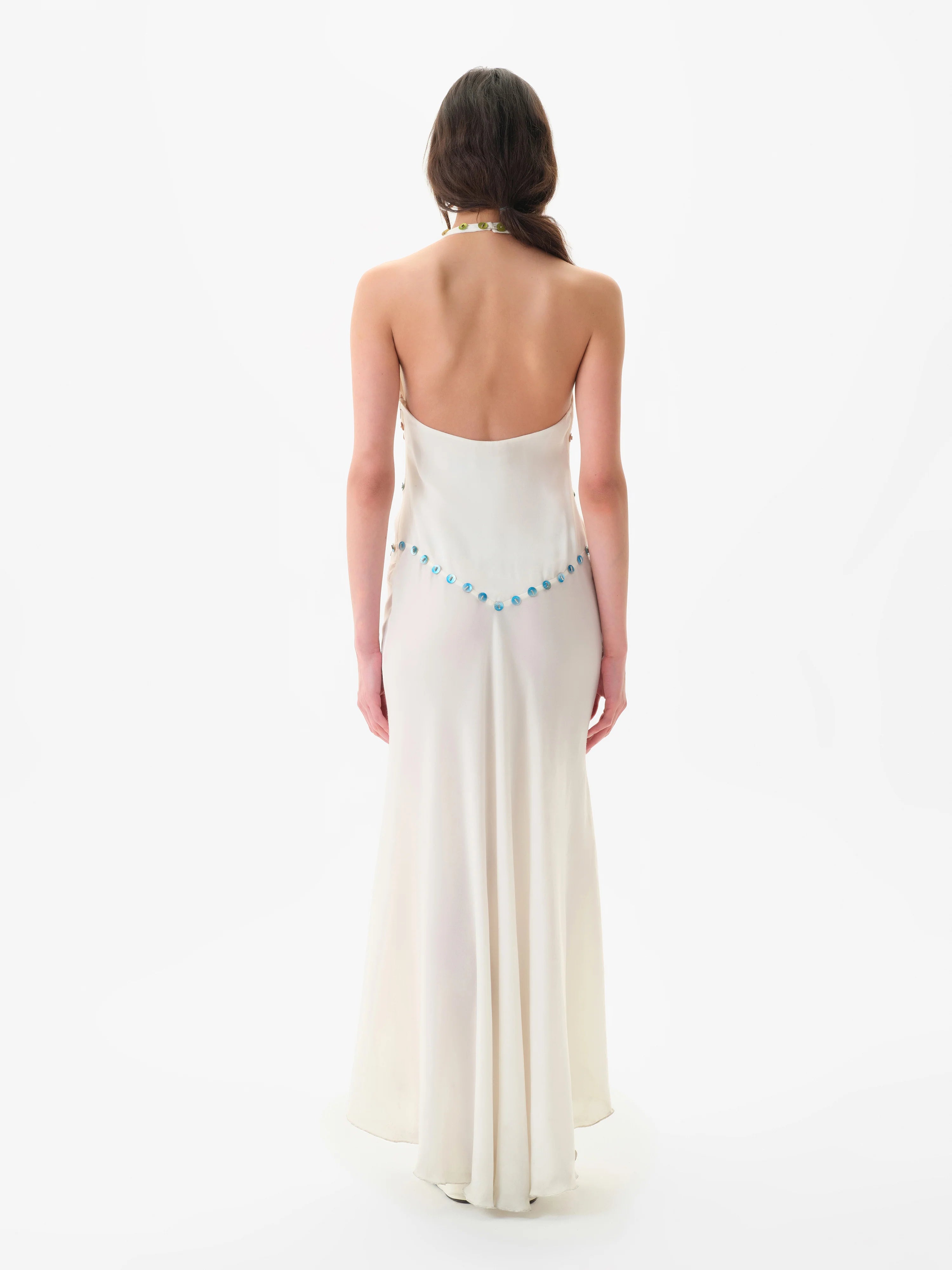 A woman with long brown hair stands facing away, wearing the Vicka Dress—a backless, floor-length white gown featuring a flared hemline and a beaded blue accent along the lower back. Elegant bead embroidery elevates the minimalist design. The background is plain white.