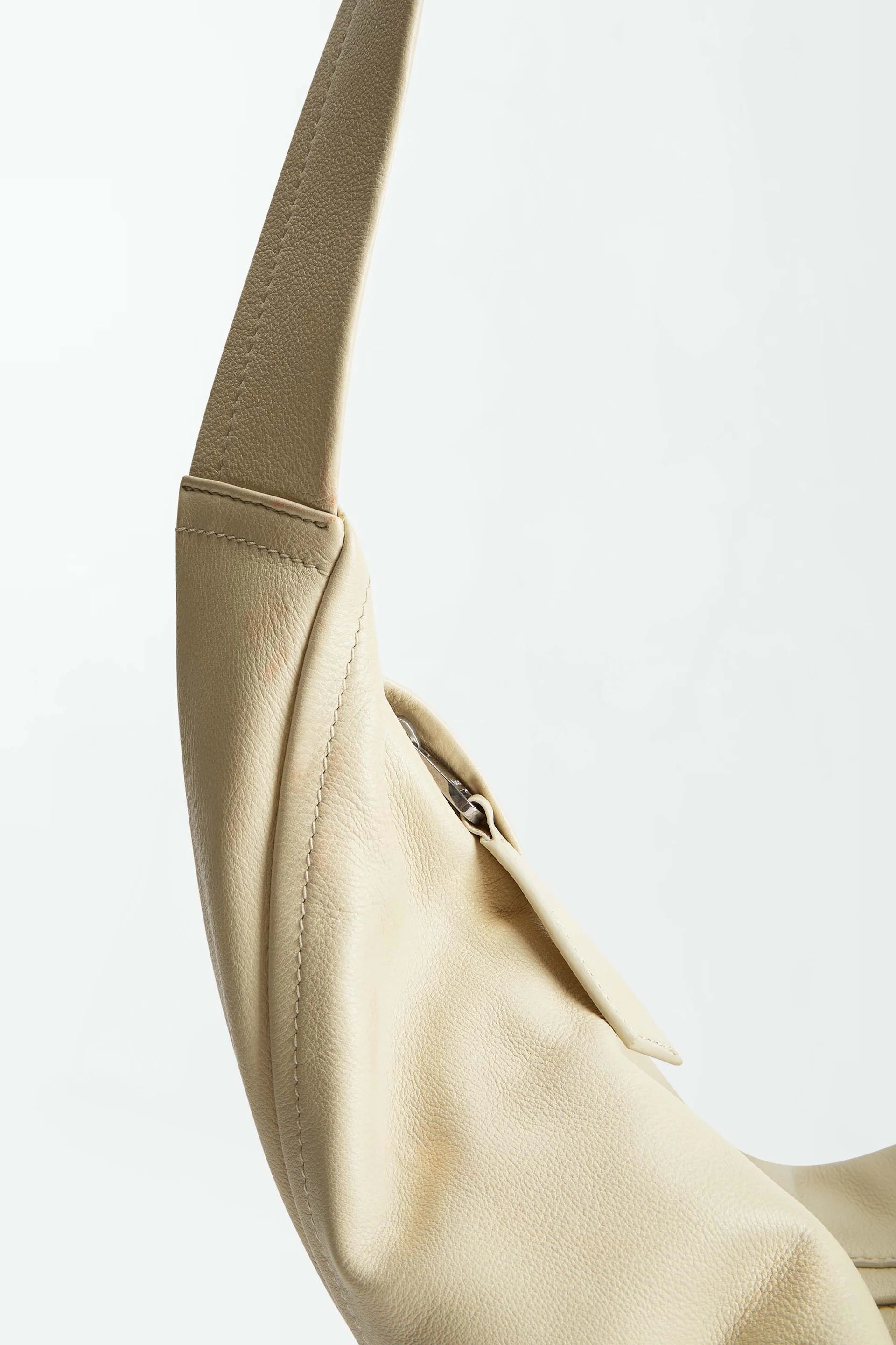 Close-up of the Vancouver Sling Bag, a beige sculptural shoulder bag crafted from supple nappa leather, featuring a curved strap and a visible seam, photographed against a plain white background.