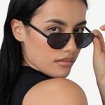 A person with long dark hair wears a sleeveless black top and tilts their head, looking over the Victoire aviator sunglasses with UV400 protection while holding the frame with one hand against a plain background.